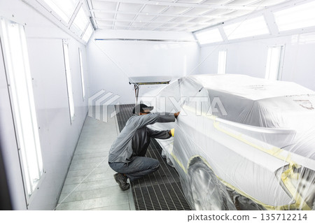 African American man technician kneeling in paint booth applying plastic film using yellow tape 135712214