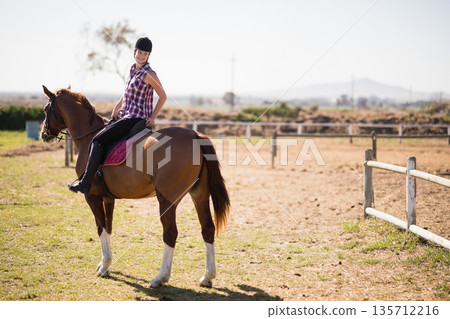 Female rider sitting astride chestnut horse in dirt paddock wearing helmet with burgundy saddle pad Female rider sitting astride chestnut horse in dirt paddock wearing helmet with burgundy saddle pad 135712216
