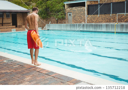 Male lifeguard holding orange rescue buoy standing barefoot at poolside brick deck, copy space 135712218