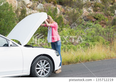 Woman inspecting engine compartment under raised hood of white sedan beside rural paved road Woman inspecting engine compartment under raised hood of white sedan beside rural paved road 135712219