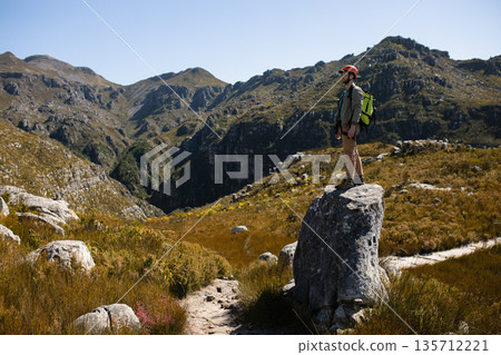 Male hiker standing atop rock on mountain trail with red helmet and green backpack, copy space 135712221
