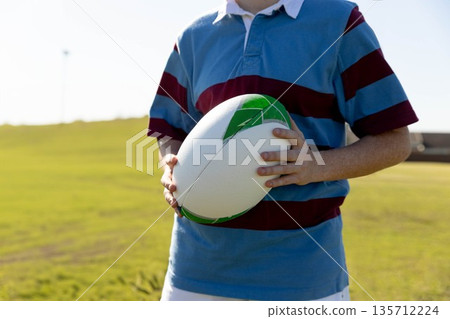 Teenage female athlete in striped rugby jersey holding white and green rugby ball on grassy field 135712224