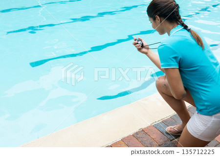 Female swim coach kneeling on outdoor pool deck holding stopwatch by lane markings, copy space Female swim coach kneeling on outdoor pool deck holding stopwatch by lane markings, copy space 135712225
