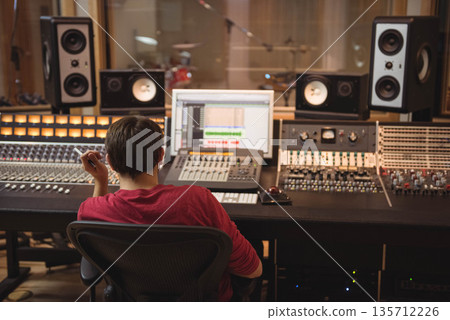 Male audio engineer mixing tracks on console in studio control room with drum kit in background 135712226