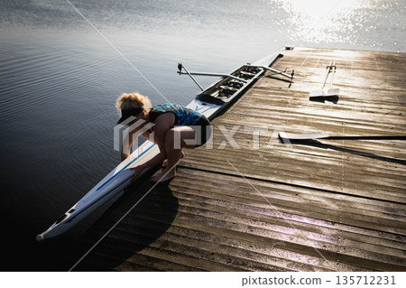 Woman wearing patterned tank top and visor guiding scull onto water at wooden dock, copy space Woman wearing patterned tank top and visor guiding scull onto water at wooden dock, copy space 135712231