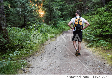 Male hiker pausing on dirt trail carrying yellow and black backpack with attached water bottle 135712232