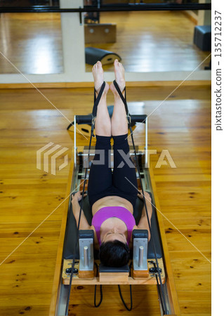 Woman in magenta sports bra and leggings performing Pilates on reformer in mirrored studio Woman in magenta sports bra and leggings performing Pilates on reformer in mirrored studio 135712237