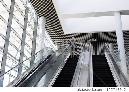 Woman riding escalator in modern lobby featuring glass wall, white columns, slatted ceiling Woman riding escalator in modern lobby featuring glass wall, white columns, slatted ceiling 135712242