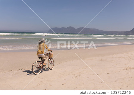 Woman riding cream cruiser bicycle on shoreline near gentle waves with wicker basket, copy space Woman riding cream cruiser bicycle on shoreline near gentle waves with wicker basket, copy space 135712246