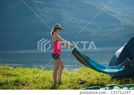 Woman standing holding tent rainfly on grassy lakeshore next to camping tent and shoes, copy space 135712247