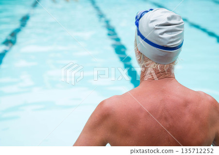 Senior man standing at pool edge with lane lines wearing swim cap and goggles, copy space 135712252