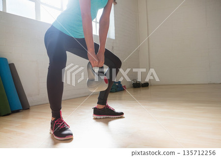 Woman wearing turquoise top and leggings doing grey kettlebell lift in gym with mats and dumbbells 135712256
