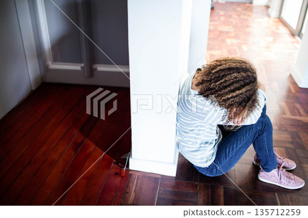 Teenage girl curling knees, resting head on arms beside white column on parquet floor in corridor 135712259