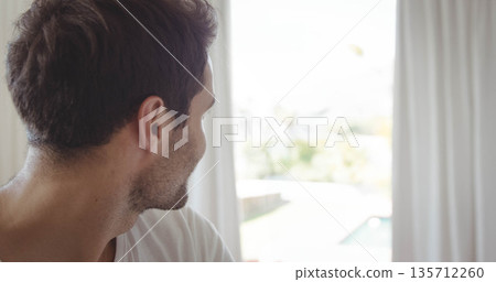 Man is wearing T-shirt and standing near glass door framed by white curtains, facing bright pool 135712260