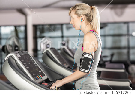 Woman running on treadmill in gym wearing sportswear and using smartphone armband, copy space Woman running on treadmill in gym wearing sportswear and using smartphone armband, copy space 135712263