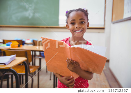 African American child smiling and holding open orange folder while standing in primary classroom 135712267
