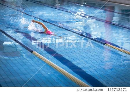 Female swimmer wearing swimsuit, pink swim cap and goggles gliding in pool lane with divider ropes Female swimmer wearing swimsuit, pink swim cap and goggles gliding in pool lane with divider ropes 135712271