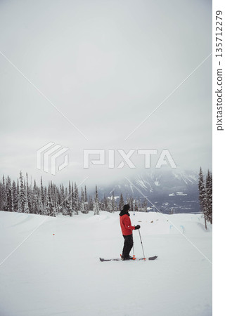 Male skier in red jacket standing on groomed slope by pines holding ski equipment, copy space Male skier in red jacket standing on groomed slope by pines holding ski equipment, copy space 135712279