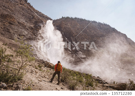 Male hiker standing on slope at waterfall in orange jacket and carrying yellow backpack, copy space Male hiker standing on slope at waterfall in orange jacket and carrying yellow backpack, copy space 135712280