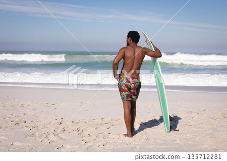 African American man standing shirtless on beach holding surfboard facing rolling ocean waves 135712281