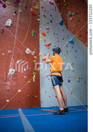 Adult male climber standing in gym wearing harness with ropes facing climbing wall, copy space 135712285