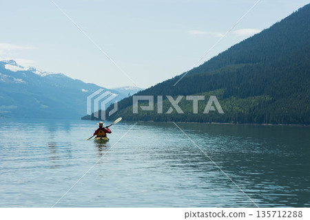 Senior male kayaker wearing hat and life vest paddling yellow kayak across mountain lake 135712288