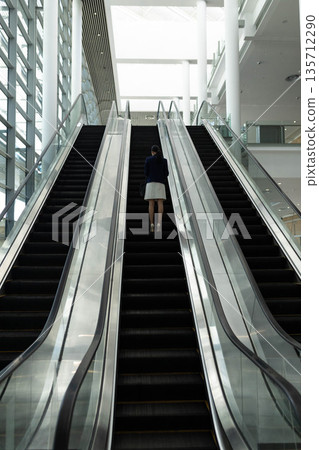 Asian businesswoman riding up escalator in modern atrium holding handbag by glass railings Asian businesswoman riding up escalator in modern atrium holding handbag by glass railings 135712290