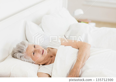 Senior woman lying on bed in bedroom gazing upward with white duvet and nightstand lamp Senior woman lying on bed in bedroom gazing upward with white duvet and nightstand lamp 135712300