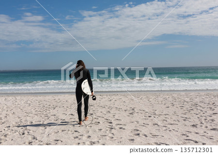 Female surfer in black wetsuit walking on beach carrying white surfboard and leash toward waves Female surfer in black wetsuit walking on beach carrying white surfboard and leash toward waves 135712301