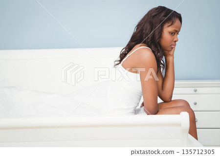 African American woman sitting in white sleepwear on edge of bed in blue bedroom, copy space African American woman sitting in white sleepwear on edge of bed in blue bedroom, copy space 135712305