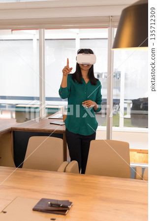 Asian woman wearing VR headset and blouse in conference room behind table reaching toward windows 135712309