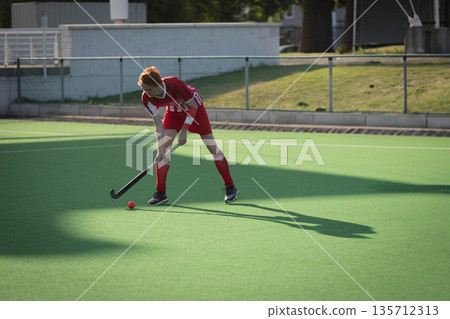 Male field hockey player bending forward, gripping stick, focusing on red ball on turf Male field hockey player bending forward, gripping stick, focusing on red ball on turf 135712313