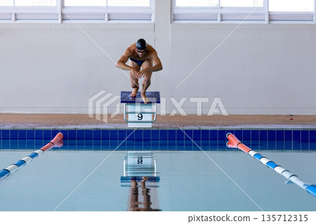 Male swimmer crouching on starting block labeled 9 at pool, wearing black swim cap and goggles Male swimmer crouching on starting block labeled 9 at pool, wearing black swim cap and goggles 135712315