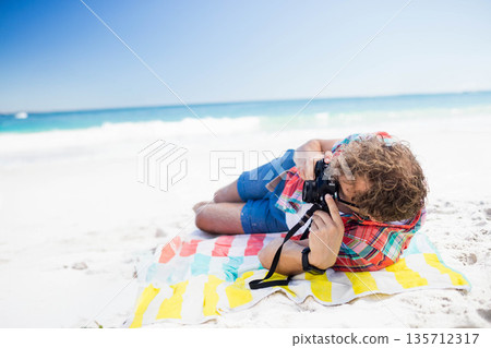 Man lying on striped beach towel in plaid shirt, aiming DSLR camera at shoreline, copy space 135712317