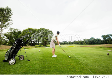 Senior female golfer holding driver while standing on tee box over golf ball and preparing drive 135712322