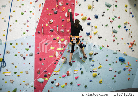 White female climber scaling colorful holds on wall at climbing gym using rope harness chalk bag White female climber scaling colorful holds on wall at climbing gym using rope harness chalk bag 135712323
