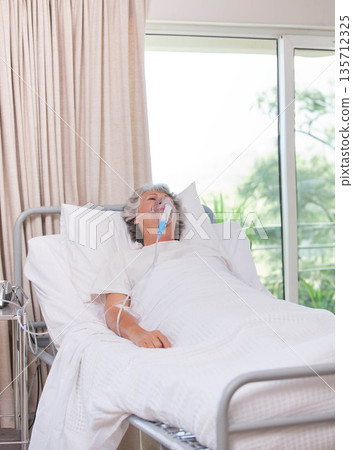 Senior female patient lying on hospital bed wearing hospital gown breathing with oxygen mask tubing Senior female patient lying on hospital bed wearing hospital gown breathing with oxygen mask tubing 135712325