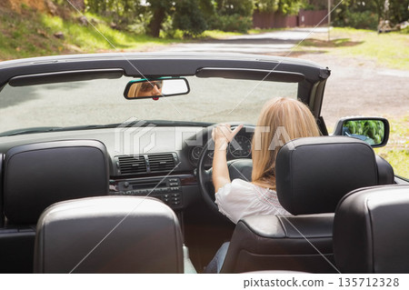 woman in mid-20s gripping steering wheel and driving convertible car on rural road under sunlight woman in mid-20s gripping steering wheel and driving convertible car on rural road under sunlight 135712328