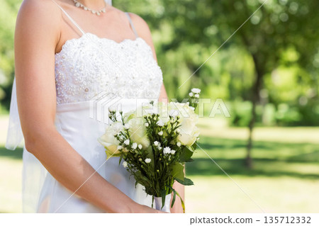 White wedding gown resting on garden lawn, holding white rose bouquet accented by greenery White wedding gown resting on garden lawn, holding white rose bouquet accented by greenery 135712332