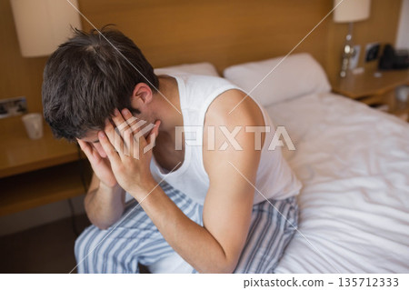 Man sitting on bed edge in bedroom with wooden headboard holding head in hands beside mug Man sitting on bed edge in bedroom with wooden headboard holding head in hands beside mug 135712333