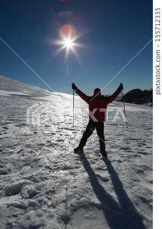 Male skier wearing red jacket celebrating on snowy mountain slope under sun with raised ski poles Male skier wearing red jacket celebrating on snowy mountain slope under sun with raised ski poles 135712335
