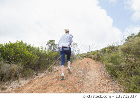 Adult female running uphill on rural dirt trail wearing gray jacket, navy leggings, running shoes 135712337