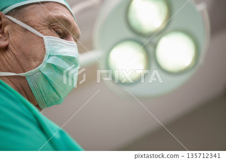 Senior male surgeon wearing green scrubs, mask, leaning under surgical light in OR copy space Senior male surgeon wearing green scrubs, mask, leaning under surgical light in OR copy space 135712341