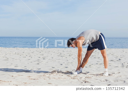 Man stretching forward in sleeveless top, sports shorts, athletic shoes on sandy beach, copy space Man stretching forward in sleeveless top, sports shorts, athletic shoes on sandy beach, copy space 135712342