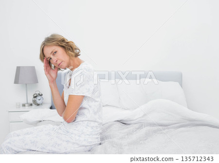 Senior woman sitting on bed in bedroom holding head with hand near nightstand lamp and clock 135712343