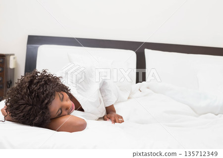 African American woman lying on side on bed at home wearing white shirt near telephone, pillows 135712349