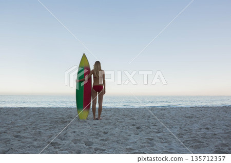 Woman standing barefoot on sandy beach at dawn facing ocean horizon and holding striped surfboard 135712357