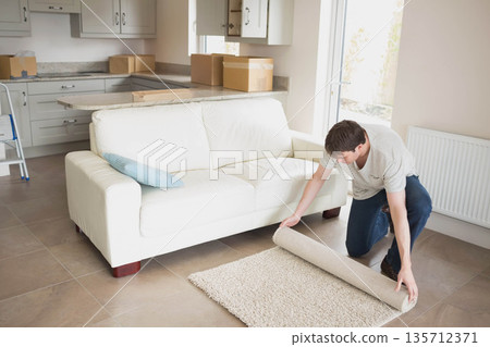 Man kneeling on tiled floor unrolling small rug beside white sofa in living room, copy space Man kneeling on tiled floor unrolling small rug beside white sofa in living room, copy space 135712371