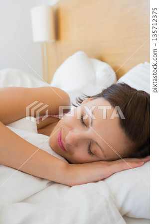 Woman sleeping on side in bedroom on white sheets pillows with wooden headboard and lamp 135712375