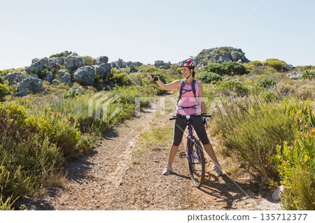 Female cyclist straddling purple mountain bike on trail with helmet and backpack holding smartphone 135712377